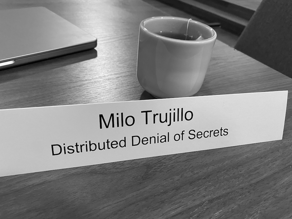A photo of a nameplate reading "Milo Trujillo - Distributed Denial of Secrets". The nameplate is on a meeting desk next to a mug of tea and someone's laptop. A photo of a nameplate reading "Milo Trujillo - Distributed Denial of Secrets". The nameplate is on a meeting desk next to a mug of tea and someone's laptop.